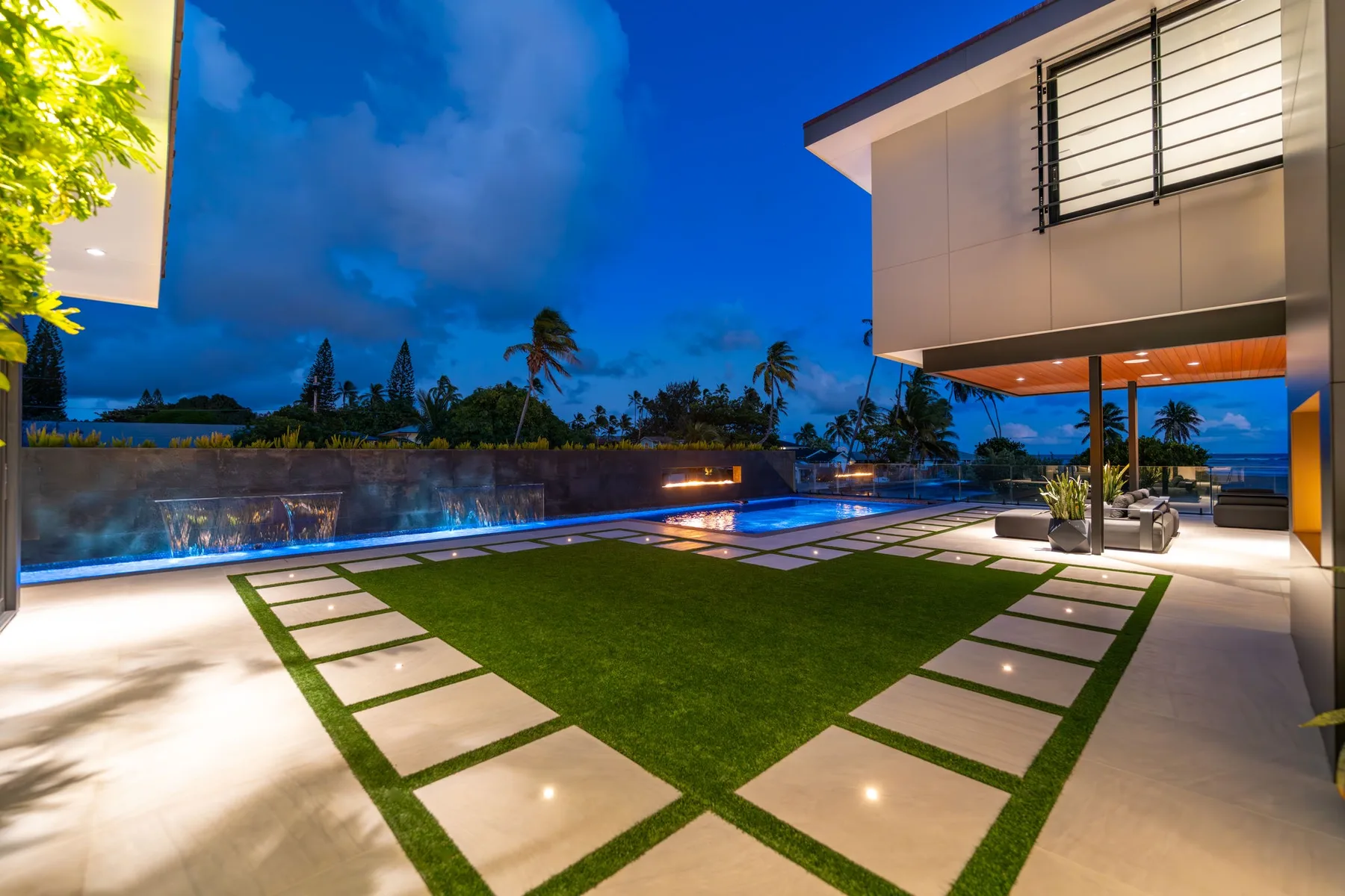 Un patio exterior moderno con césped rectangular, losas para caminar y una piscina iluminada.