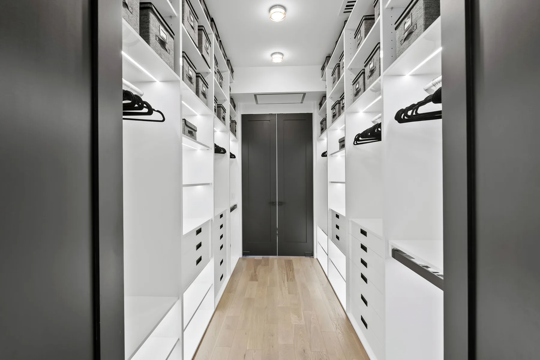 Un vestidor iluminado con estantes y cajones blancos a lo largo de un pasillo con suelo de madera.