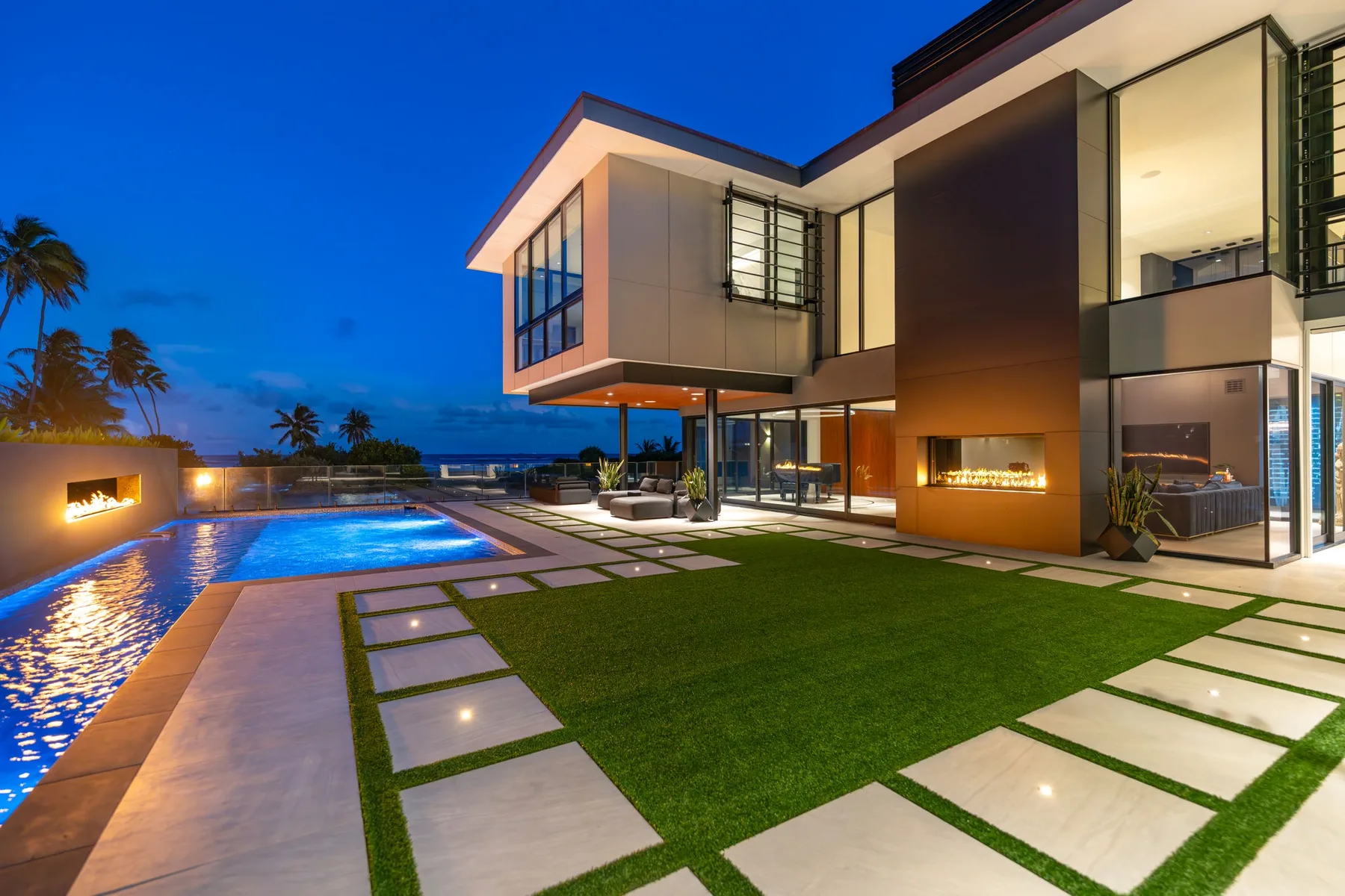 Una casa moderna y una piscina iluminada bordean un césped y un camino pavimentado.