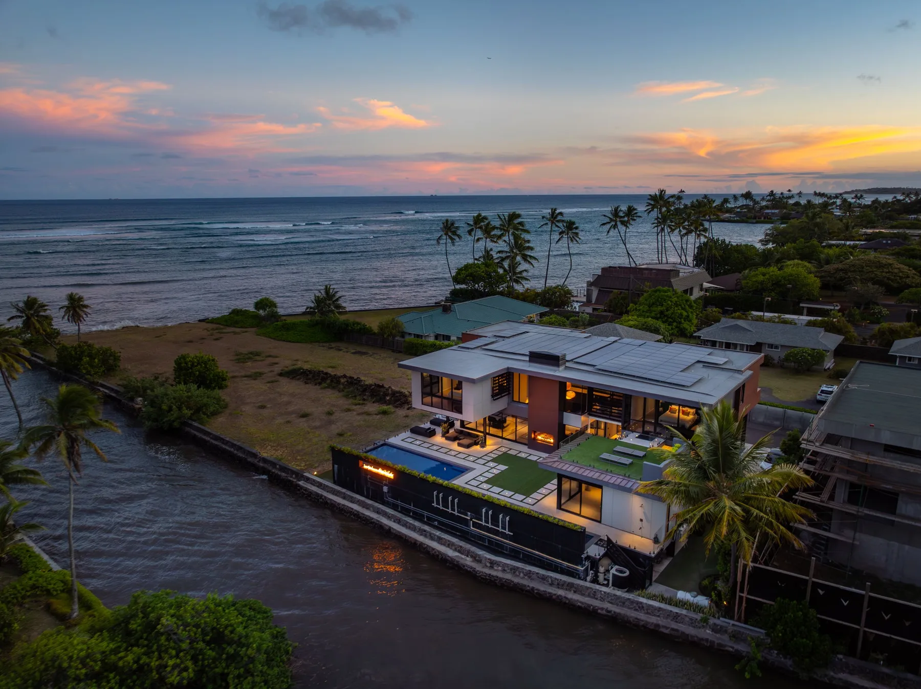 Una casa moderna junto a la costa al atardecer, con palmeras y una piscina.