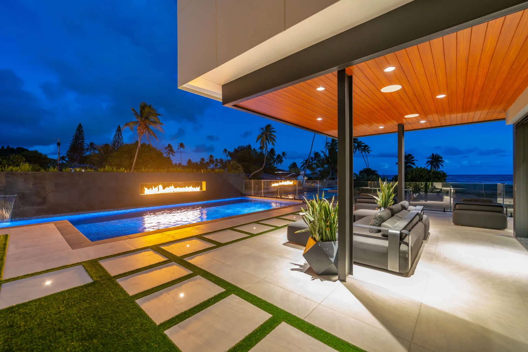 Una sala exterior moderna da a una piscina iluminada y al océano al anochecer.