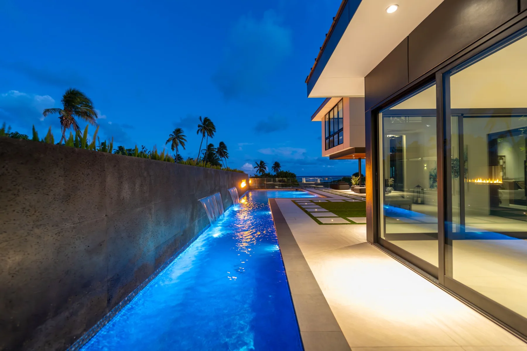 Una piscina exterior iluminada se extiende junto a un edificio moderno con fachada de vidrio al anochecer.