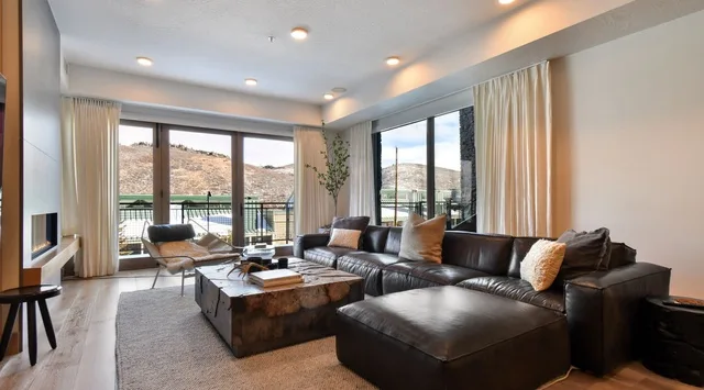 A living room with dark seating, a coffee table, and windows overlooking a hillside.
