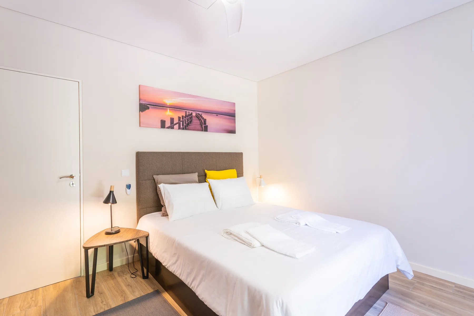 A bright bedroom with a neatly made bed and sunset artwork above the headboard.