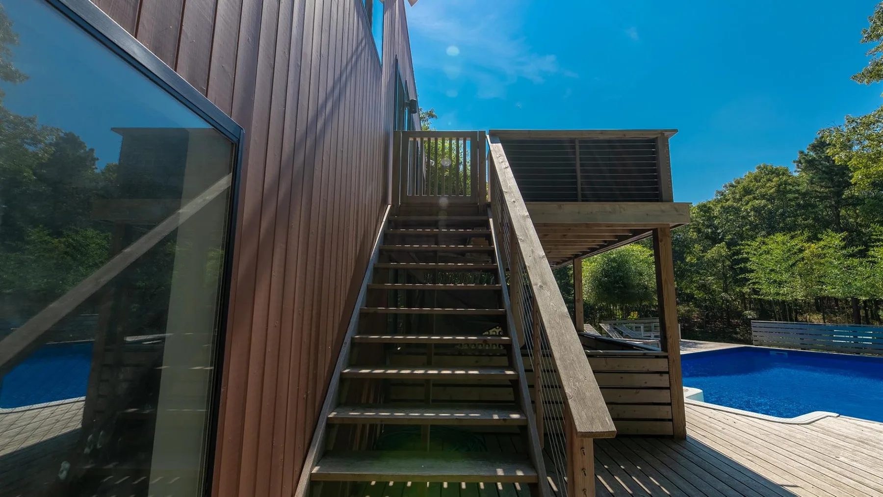 An outdoor wooden deck has stairs leading to an upper landing beside a swimming pool.
