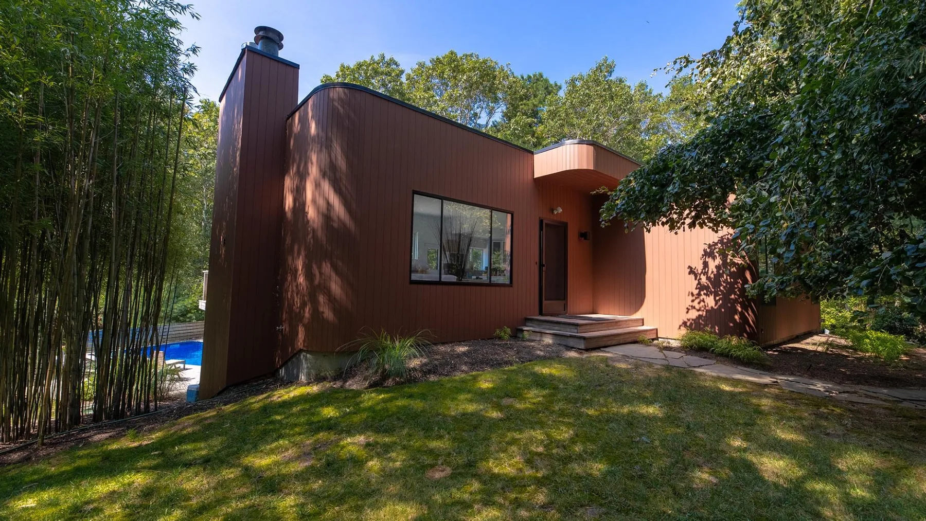 A modern brown house sits among trees beside a grassy yard.