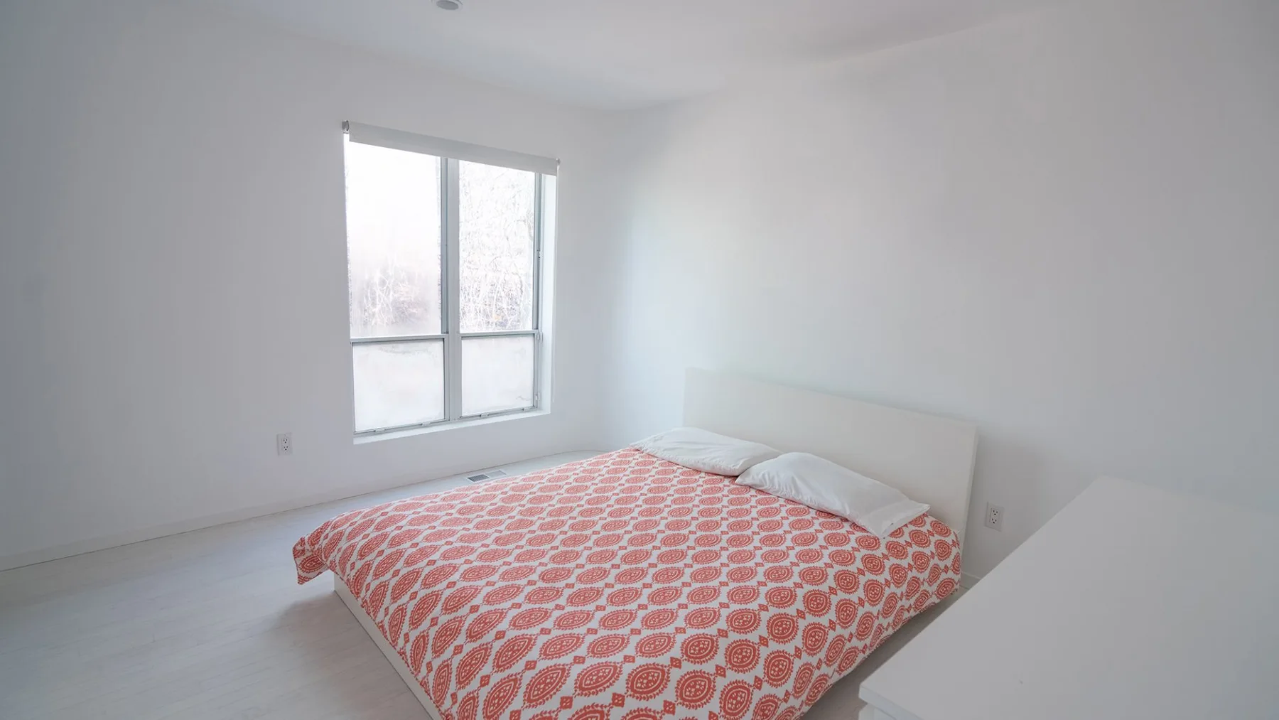 A bedroom with a bed covered in a red patterned bedspread beside a window.