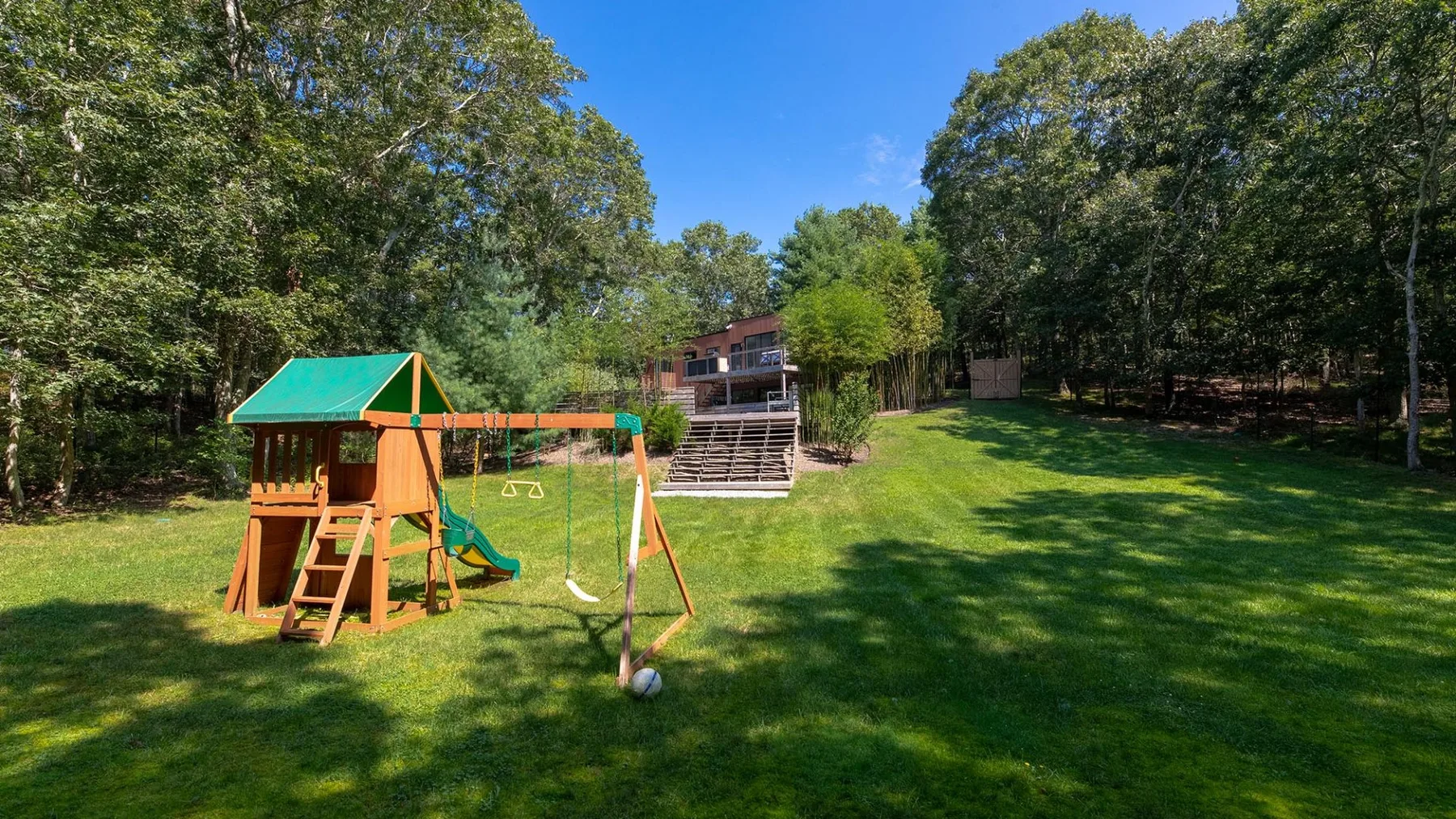 A grassy backyard features a wooden playset and a house among trees.