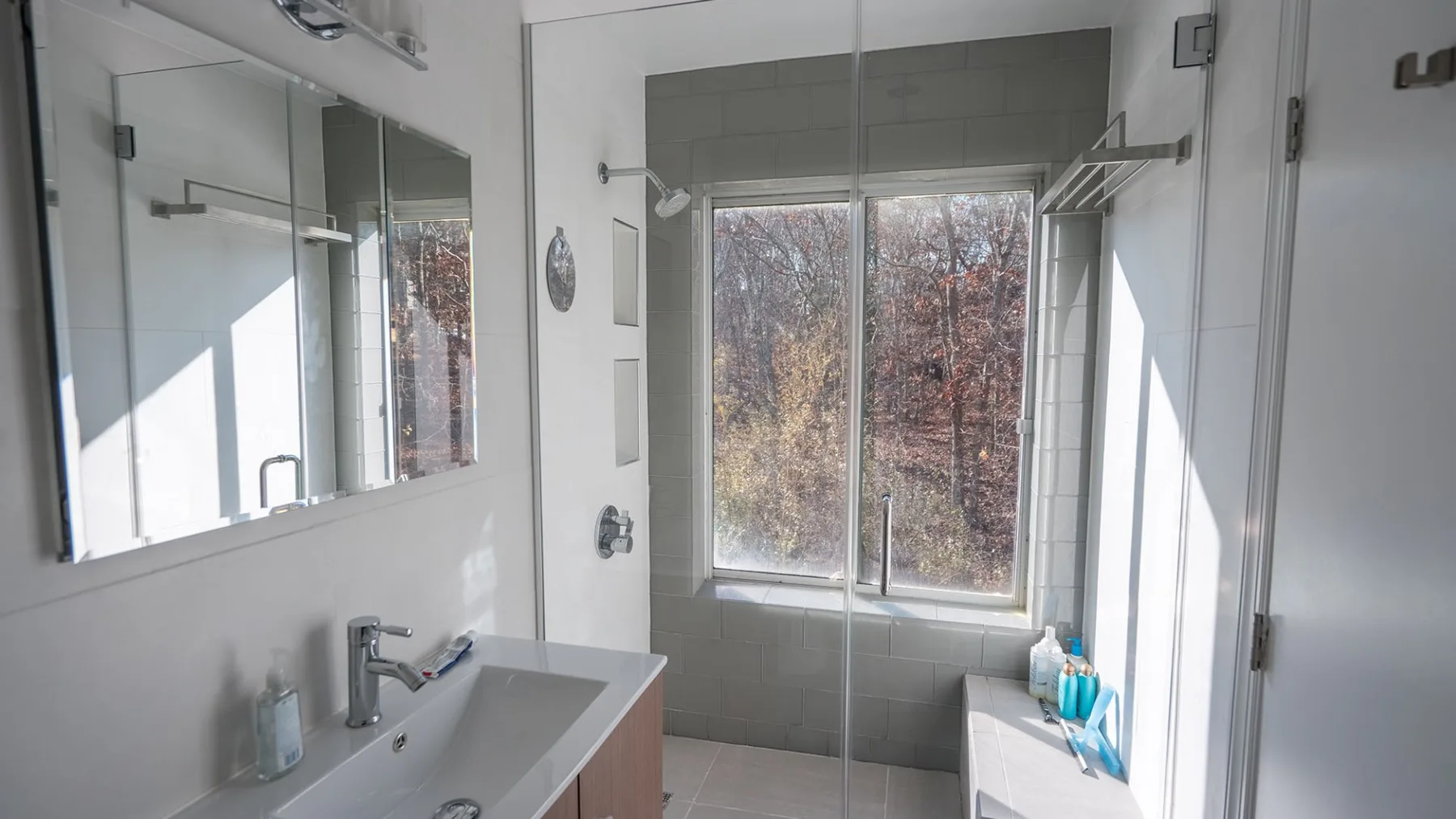 A bright bathroom with a sink, mirror, and shower beside a large window.