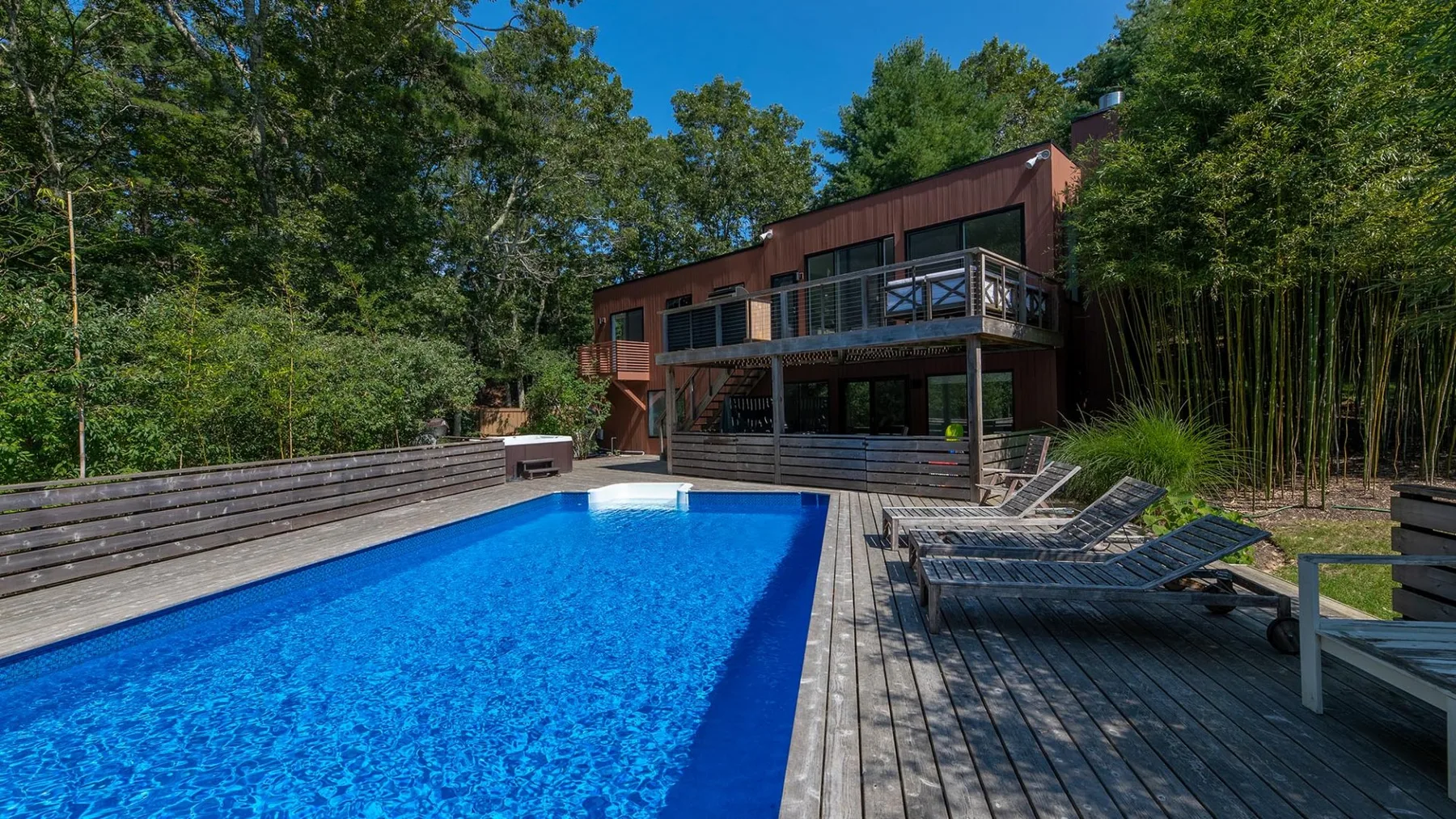 A swimming pool and wooden deck sit beside a modern house surrounded by trees.