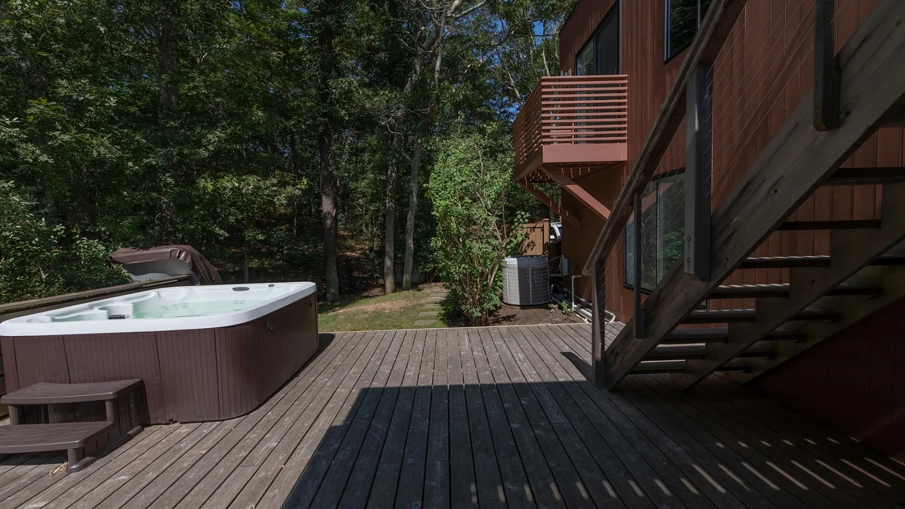A wooden deck beside a wooded area, with an outdoor hot tub and stairs.