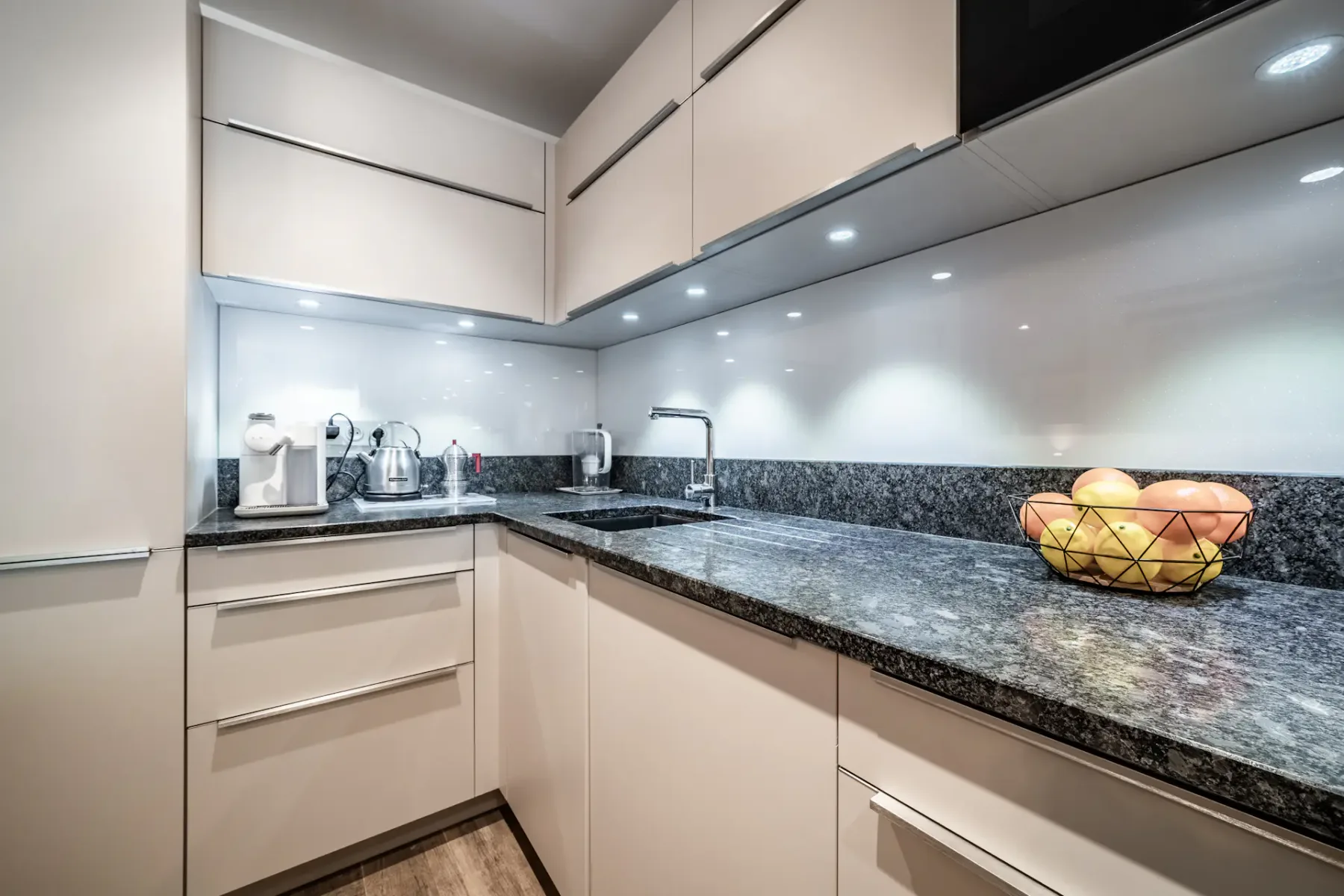Modern kitchen with white cabinets, black granite countertops, built-in appliances, wooden flooring, and overhead lighting.