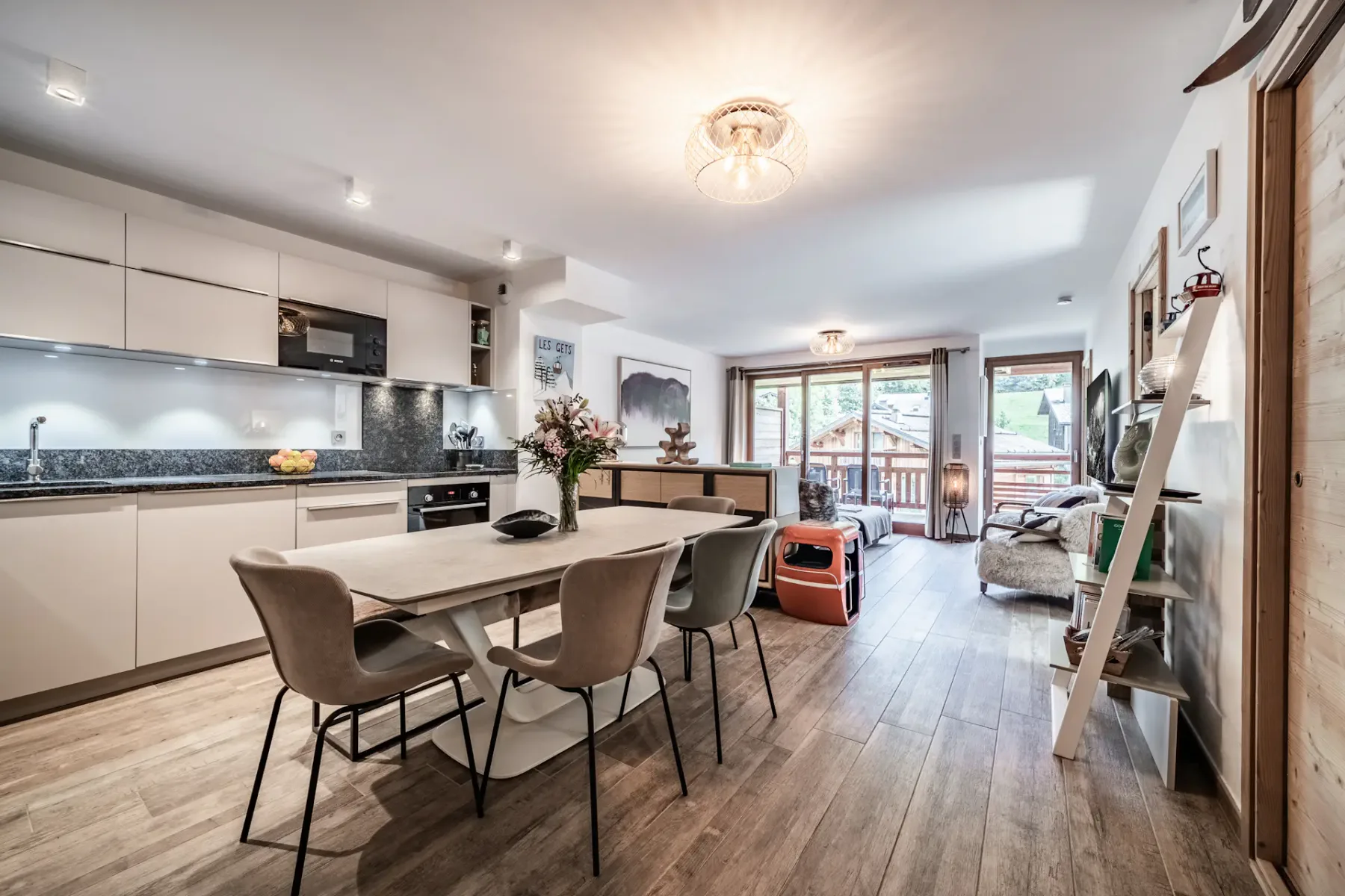Modern open-plan kitchen and dining area with wooden flooring, sleek cabinets, a table with chairs, and large windows leading to a balcony.