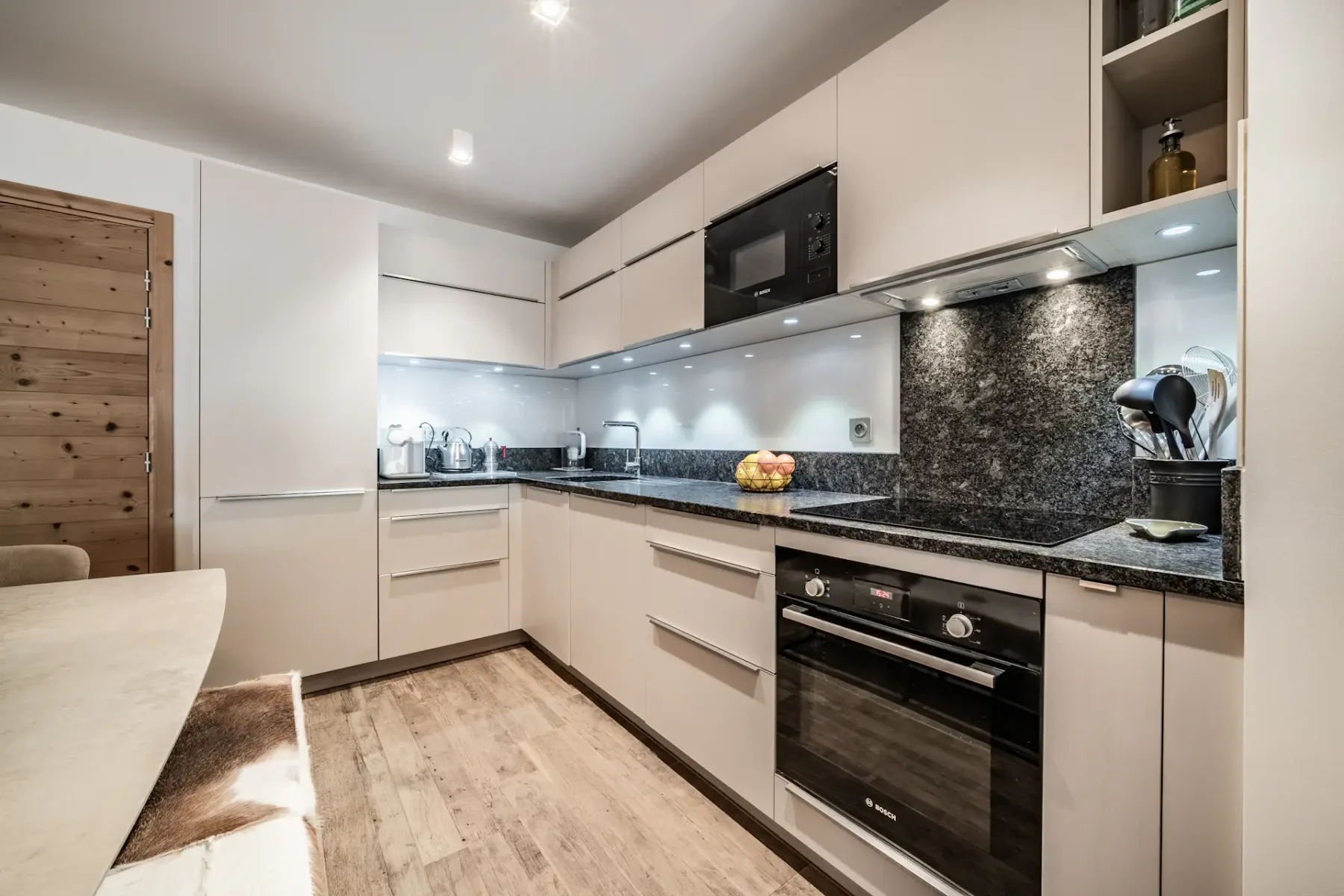 Modern kitchen with white cabinets, black granite countertops, built-in appliances, wooden flooring, and overhead lighting.