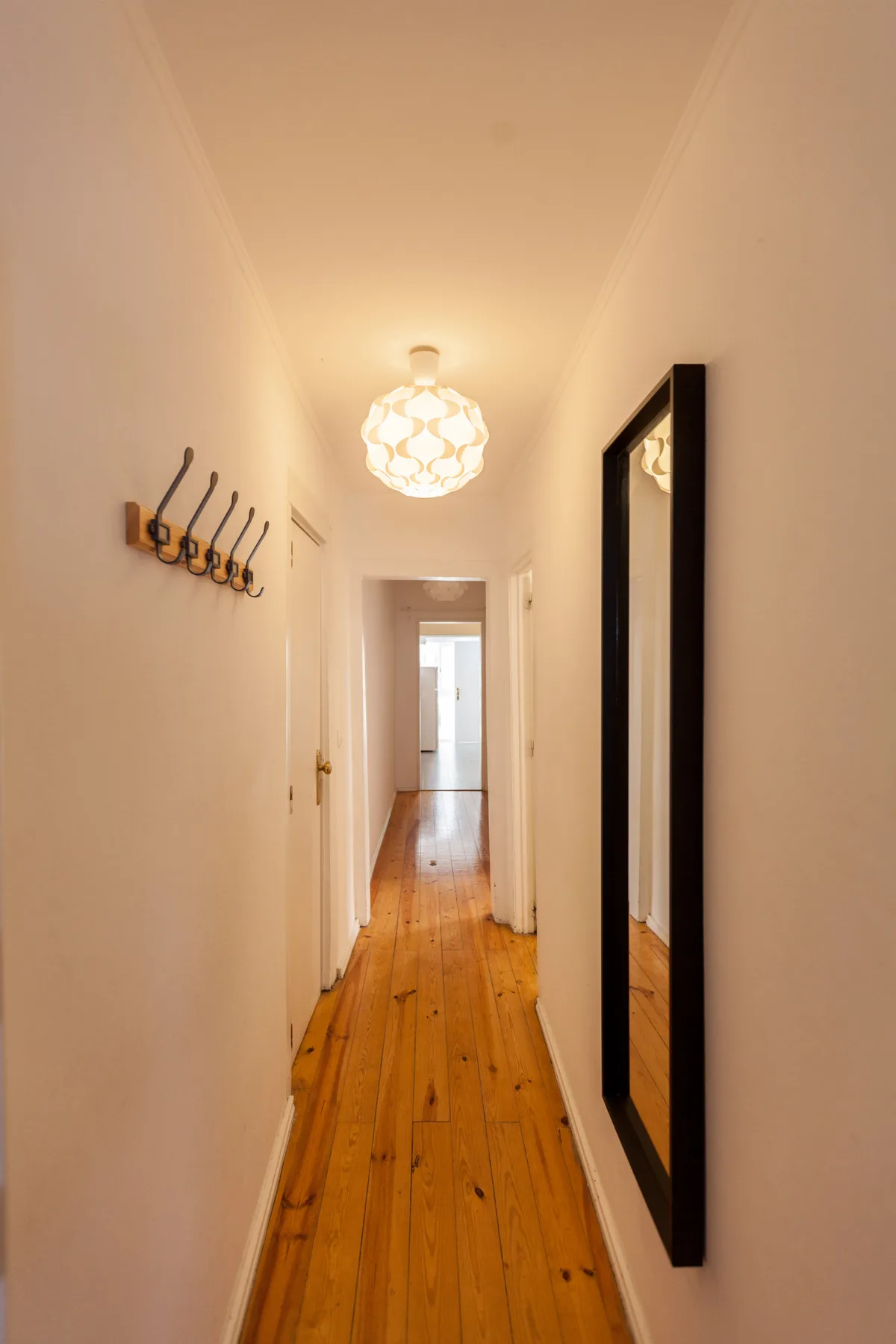 Un pasillo estrecho con suelo de madera, ganchos en la pared y una lámpara colgante.