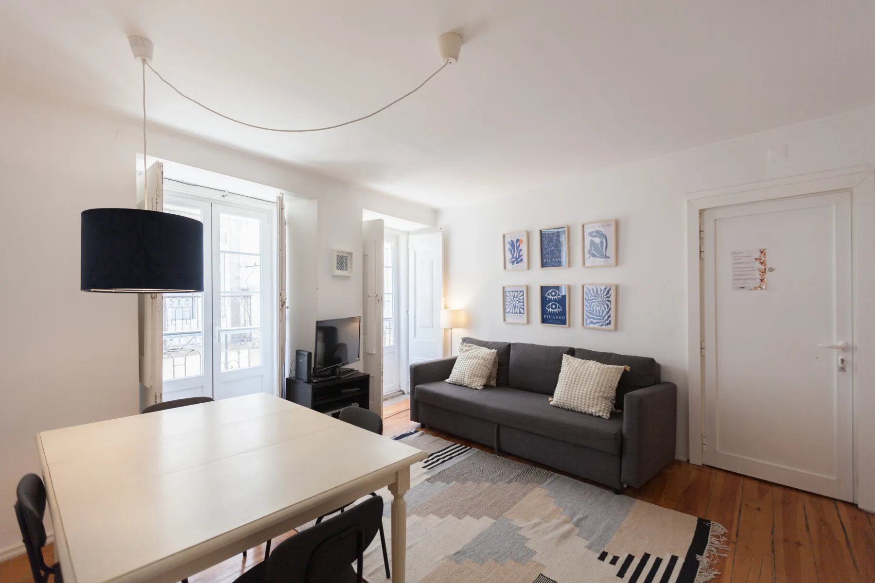 Una sala luminosa tiene un sofá oscuro, una mesa de comedor y cuadros enmarcados.