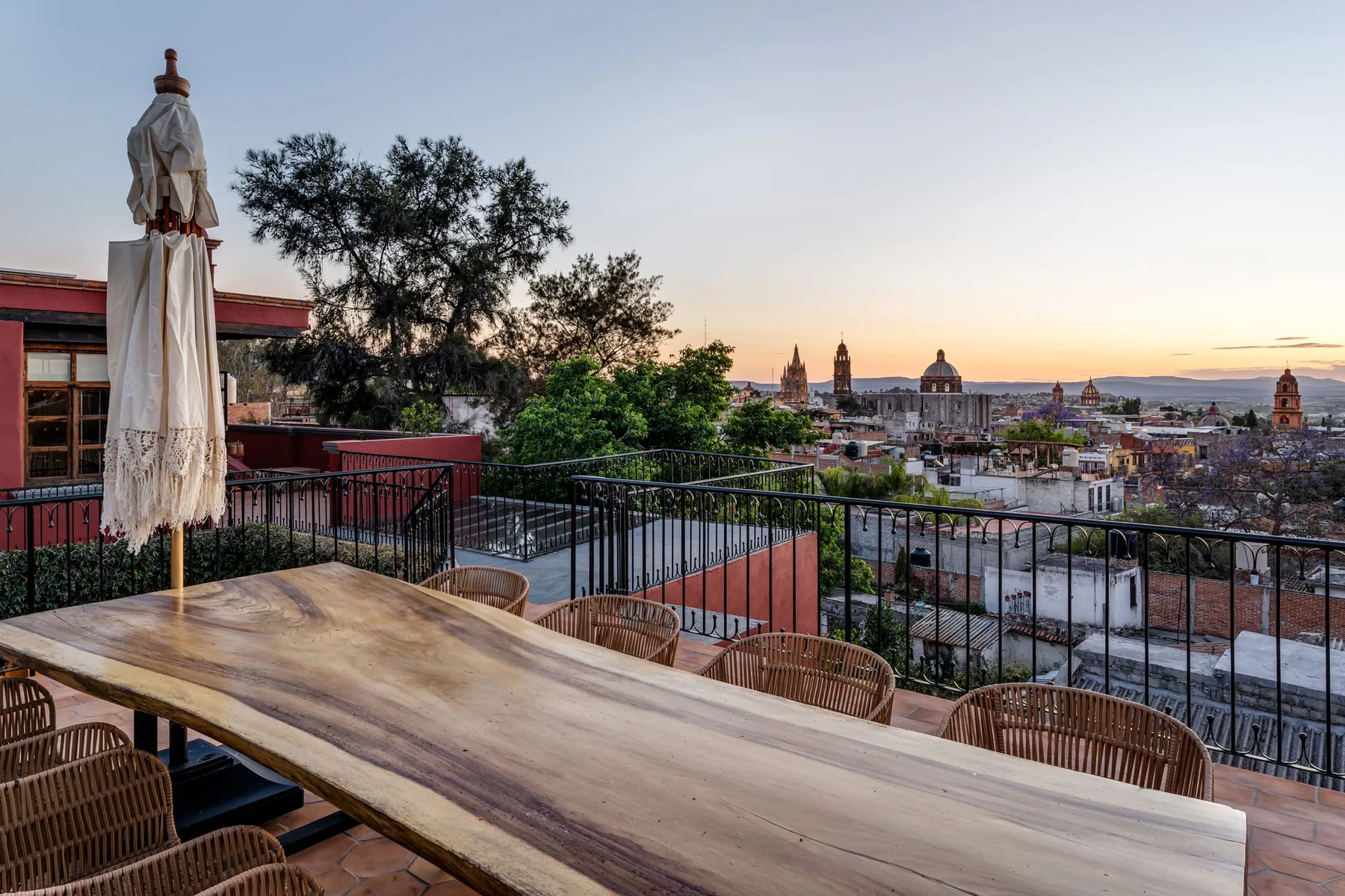 A rooftop terrace overlooks a city skyline at sunset, with an outdoor dining table.