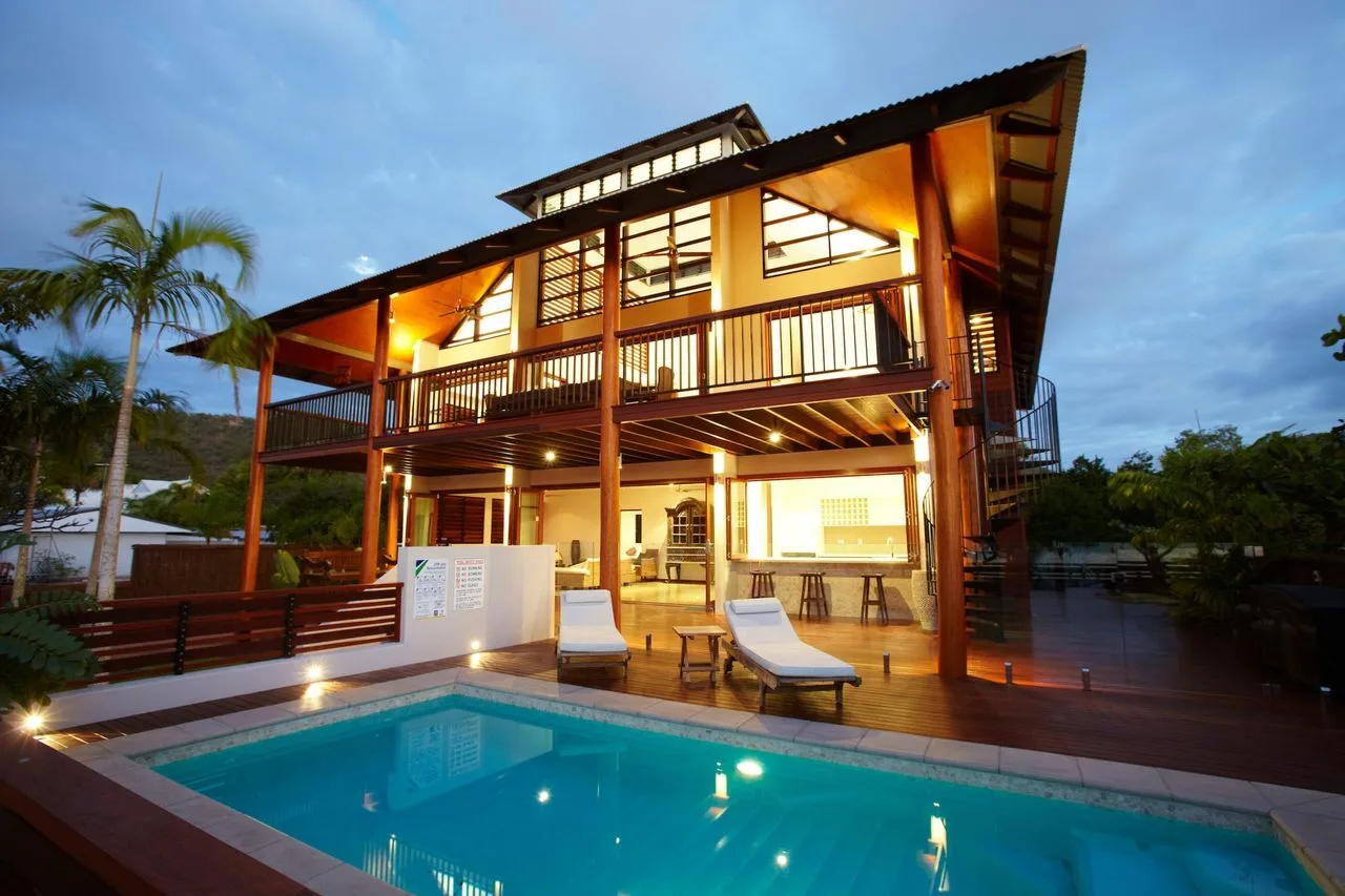 A lit two-story house overlooks a swimming pool at dusk.