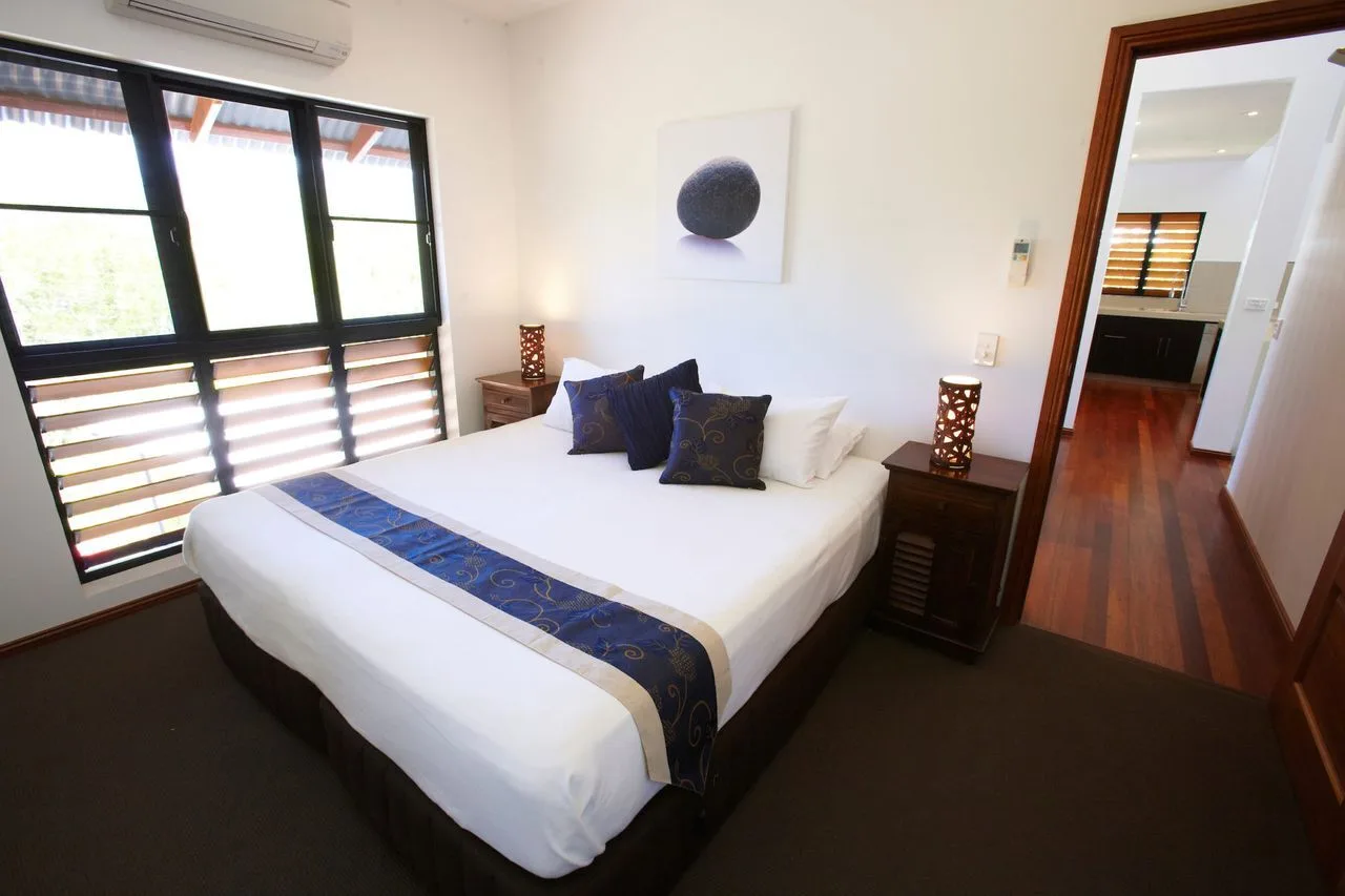 A bedroom with a neatly made bed, dark pillows, and large windows.