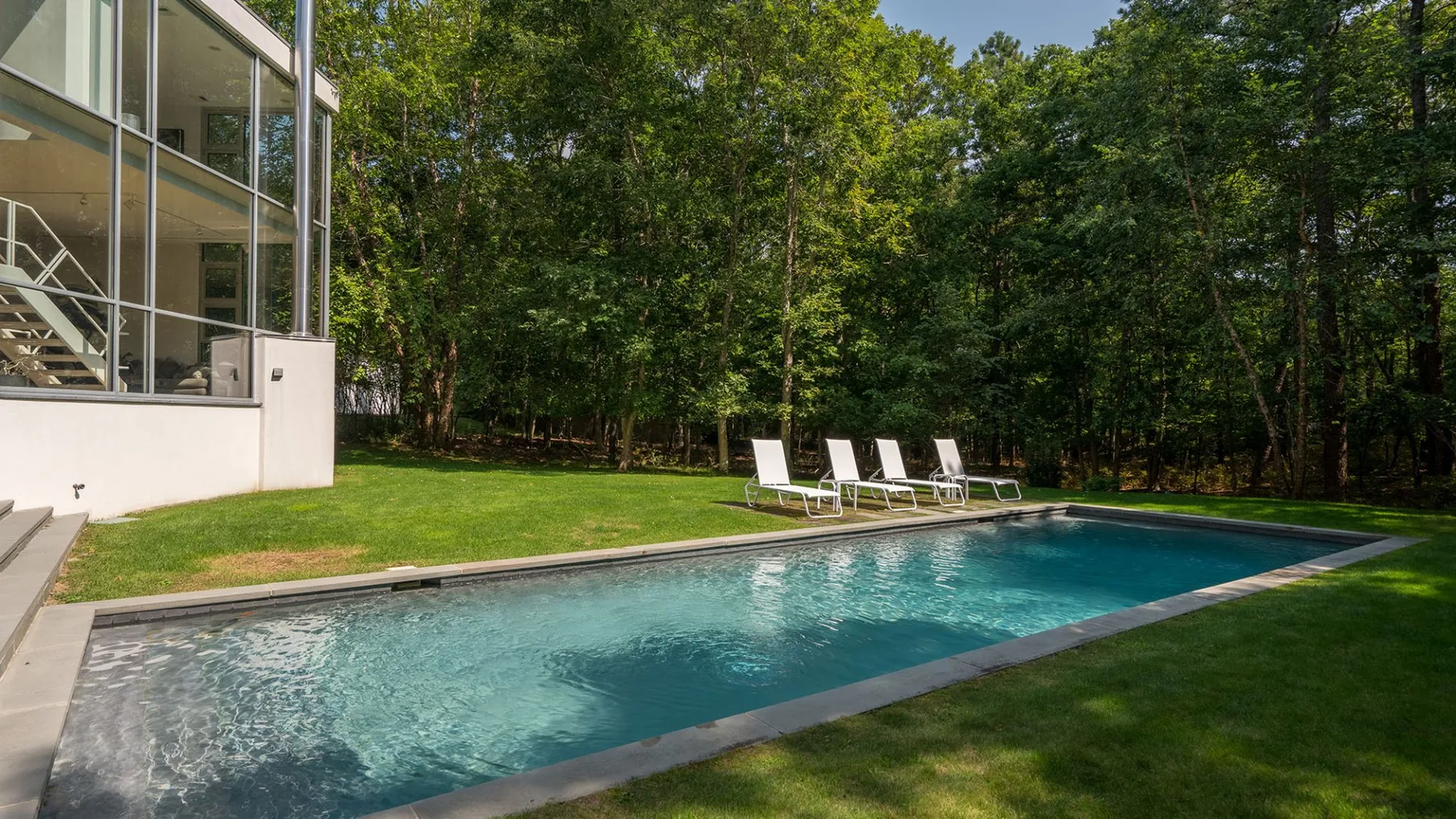 An outdoor swimming pool beside a modern glass-walled building and lawn.