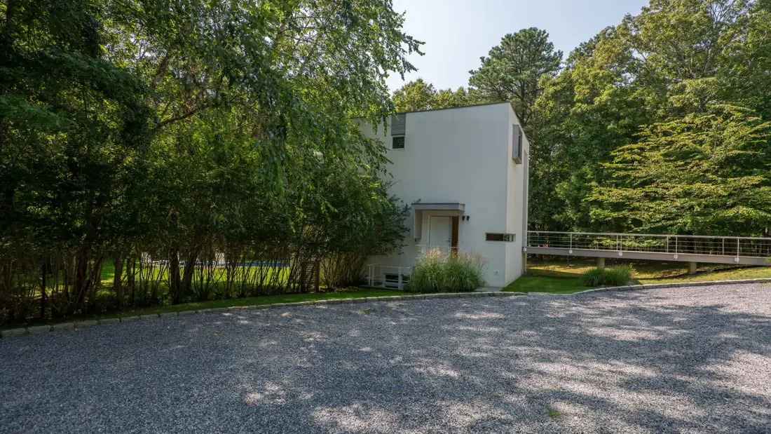 A modern building stands beside a gravel driveway, surrounded by trees.