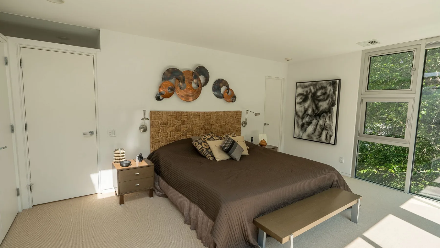 A bedroom with a bed, bedside table, wall art, and large windows.