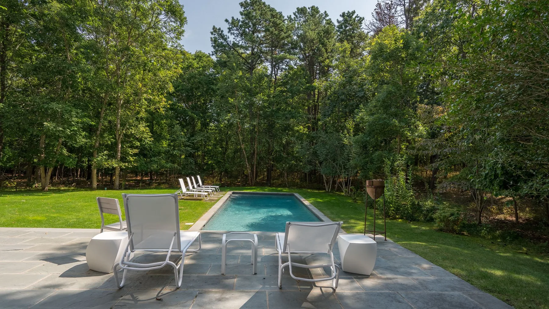 An outdoor swimming pool bordered by patio seating and a wooded yard.