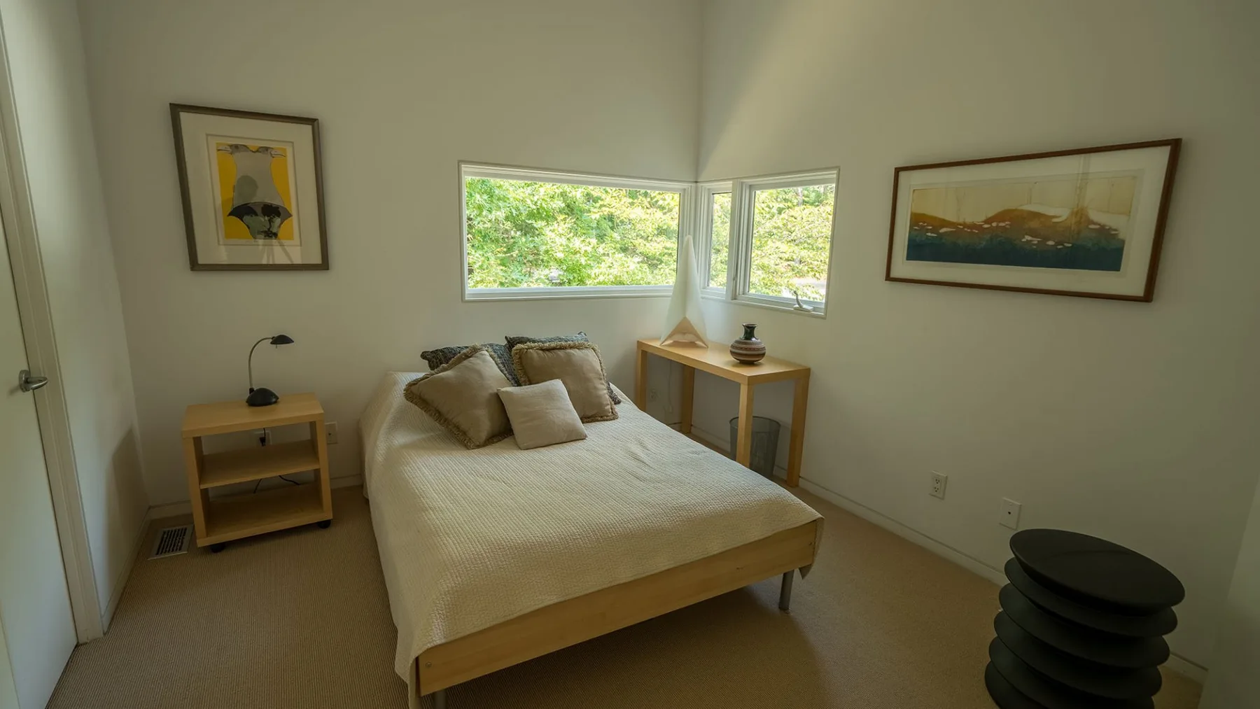 A bright bedroom with a bed beneath corner windows overlooking greenery.