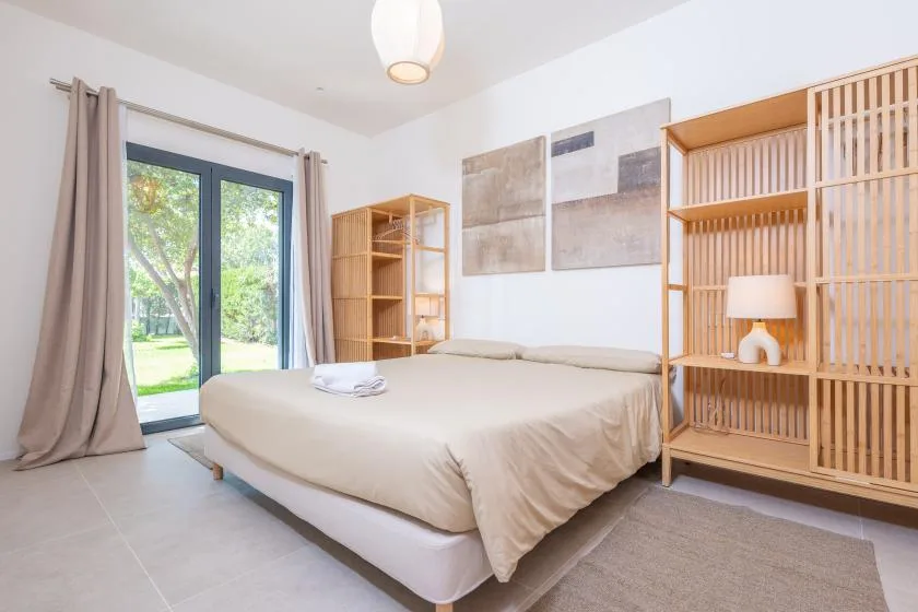 Un dormitorio luminoso con una cama, estanterías de madera y una puerta de vidrio junto a la vegetación.