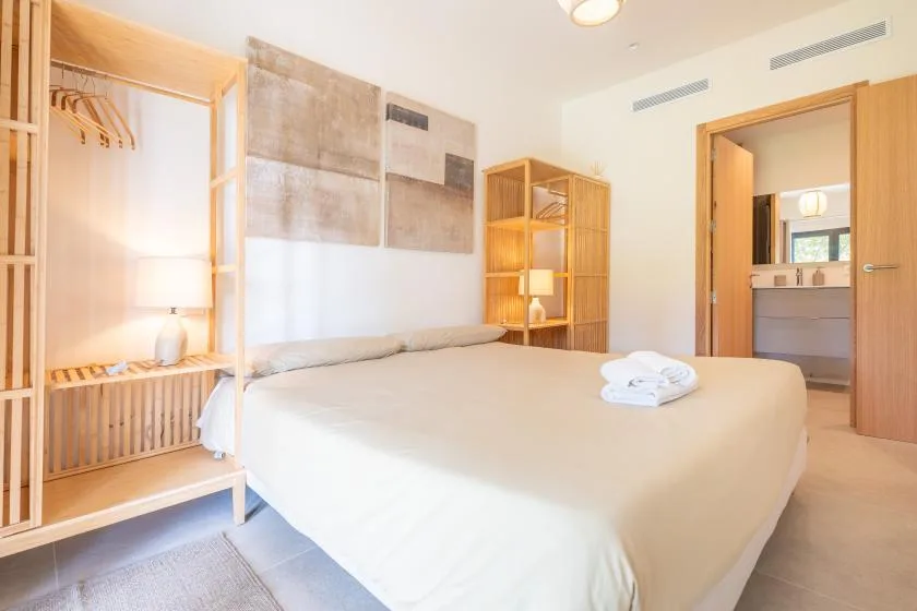 Un dormitorio luminoso con una cama, almacenamiento de madera abierto y una puerta al baño.