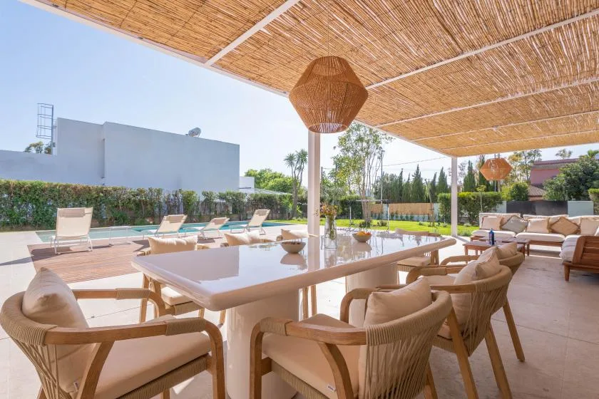 Una zona exterior de comedor y estar se encuentra bajo un toldo tejido junto a una piscina.