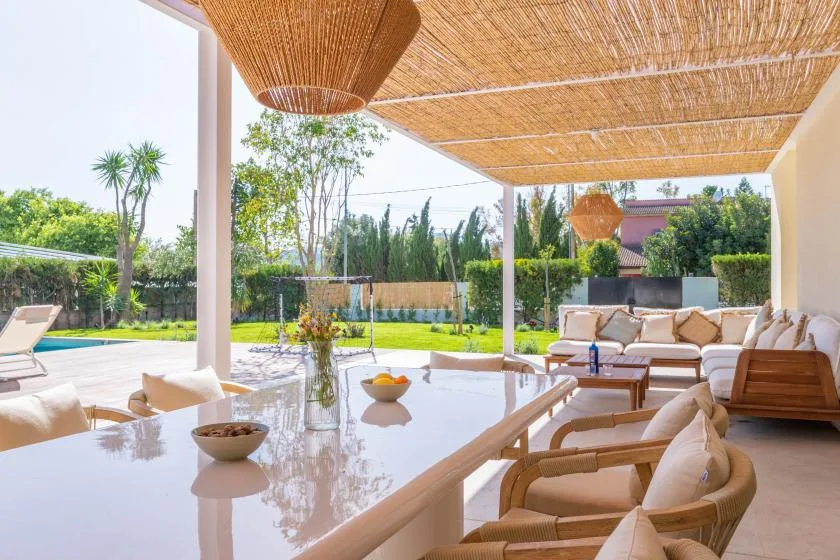 Una zona exterior de comedor y estar a la sombra da a un jardín y una piscina.