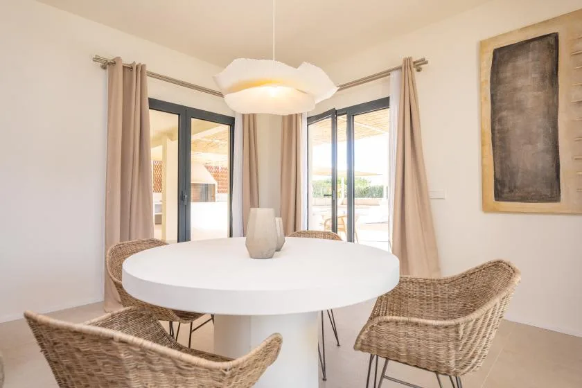 Un comedor luminoso con una mesa redonda y asientos tejidos junto a grandes ventanas.