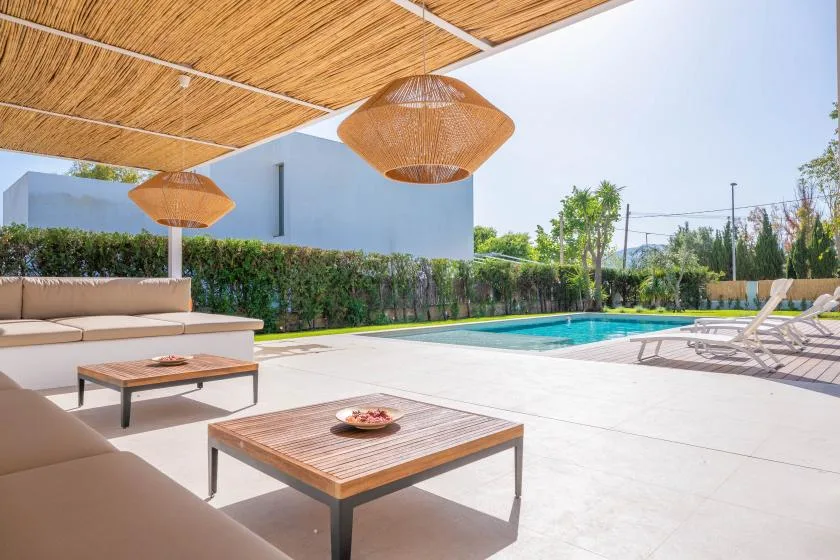 Un patio junto a la piscina con asientos acolchados, mesas bajas y toldos tejidos.