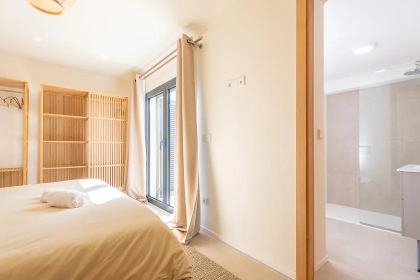 Un dormitorio luminoso con una cama junto a ventanas altas y un baño contiguo.