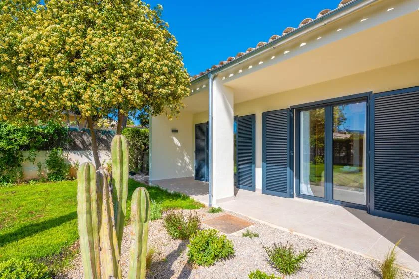 Exterior de una casa con patio sombreado, contraventanas oscuras y jardín ajardinado.