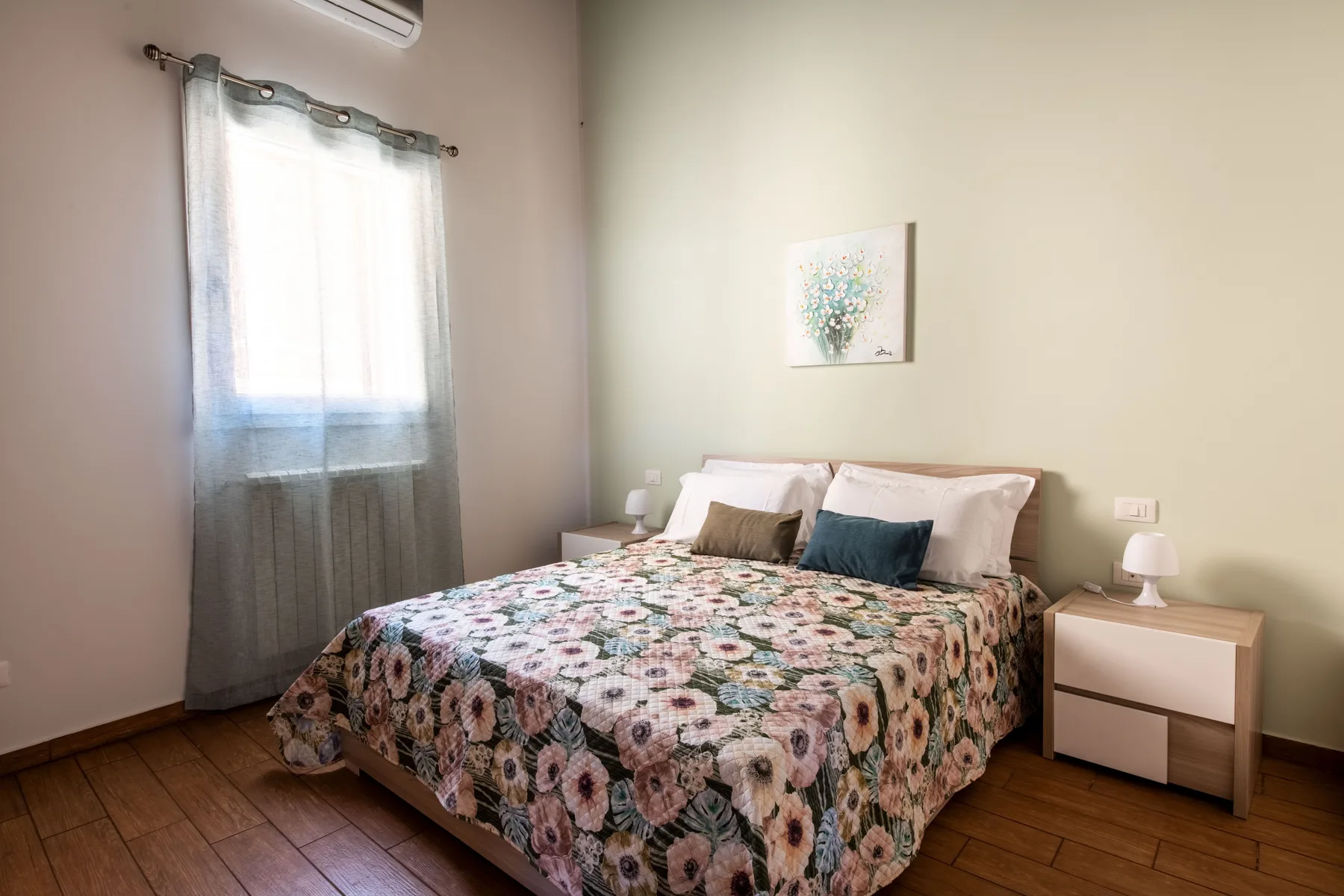 Un dormitorio con una colcha floral, mesitas de noche y una ventana con cortinas.