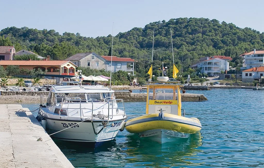 Boats are moored beside a waterfront promenade, with houses and a green hillside beyond.