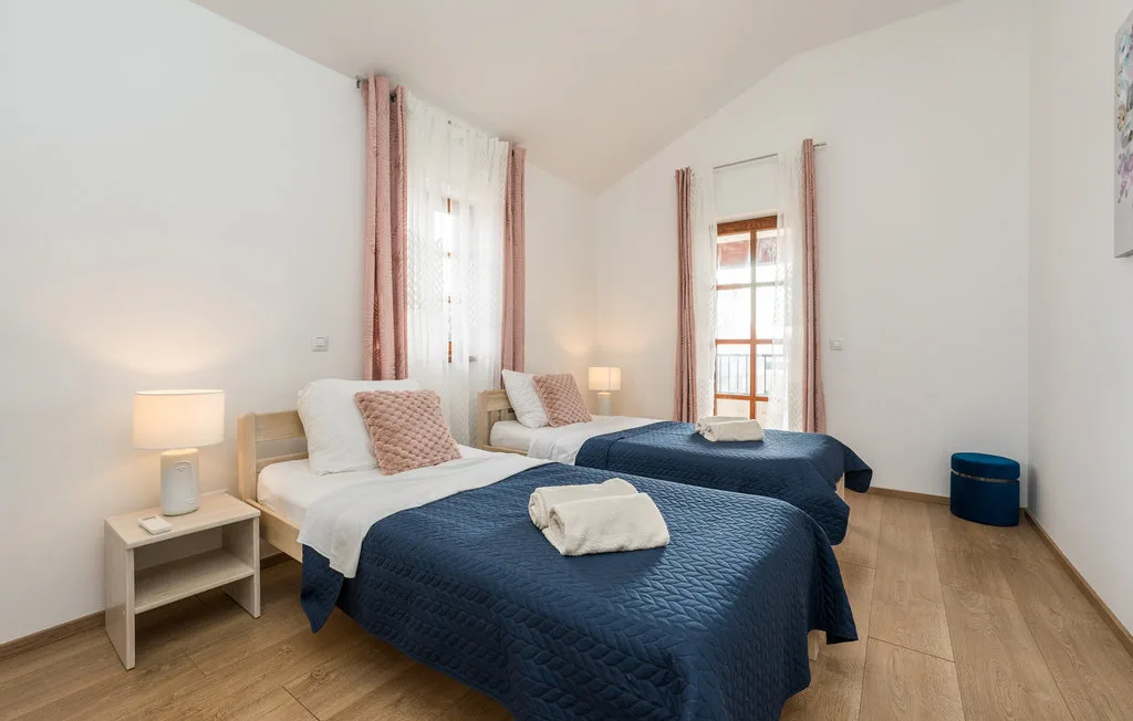 A bright bedroom with two beds dressed in blue covers and tall windows.