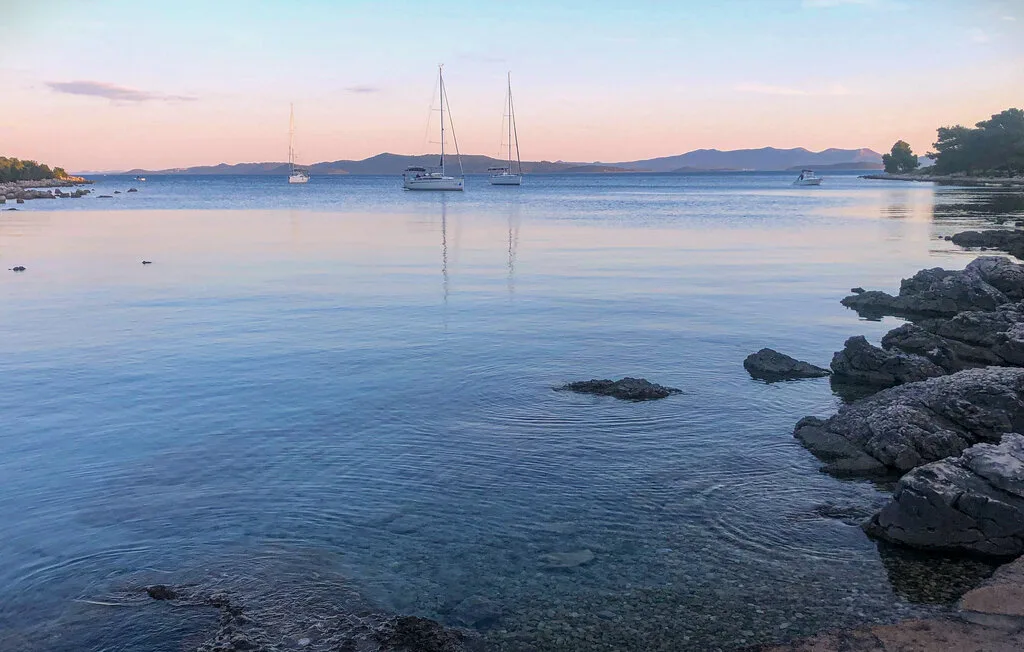 A calm coastal bay at sunset, with sailboats anchored offshore.
