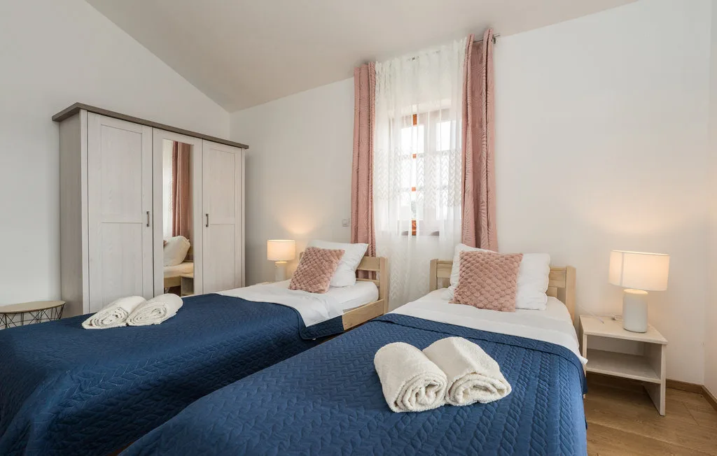 A bright bedroom with two beds dressed in blue bedding beside a window.