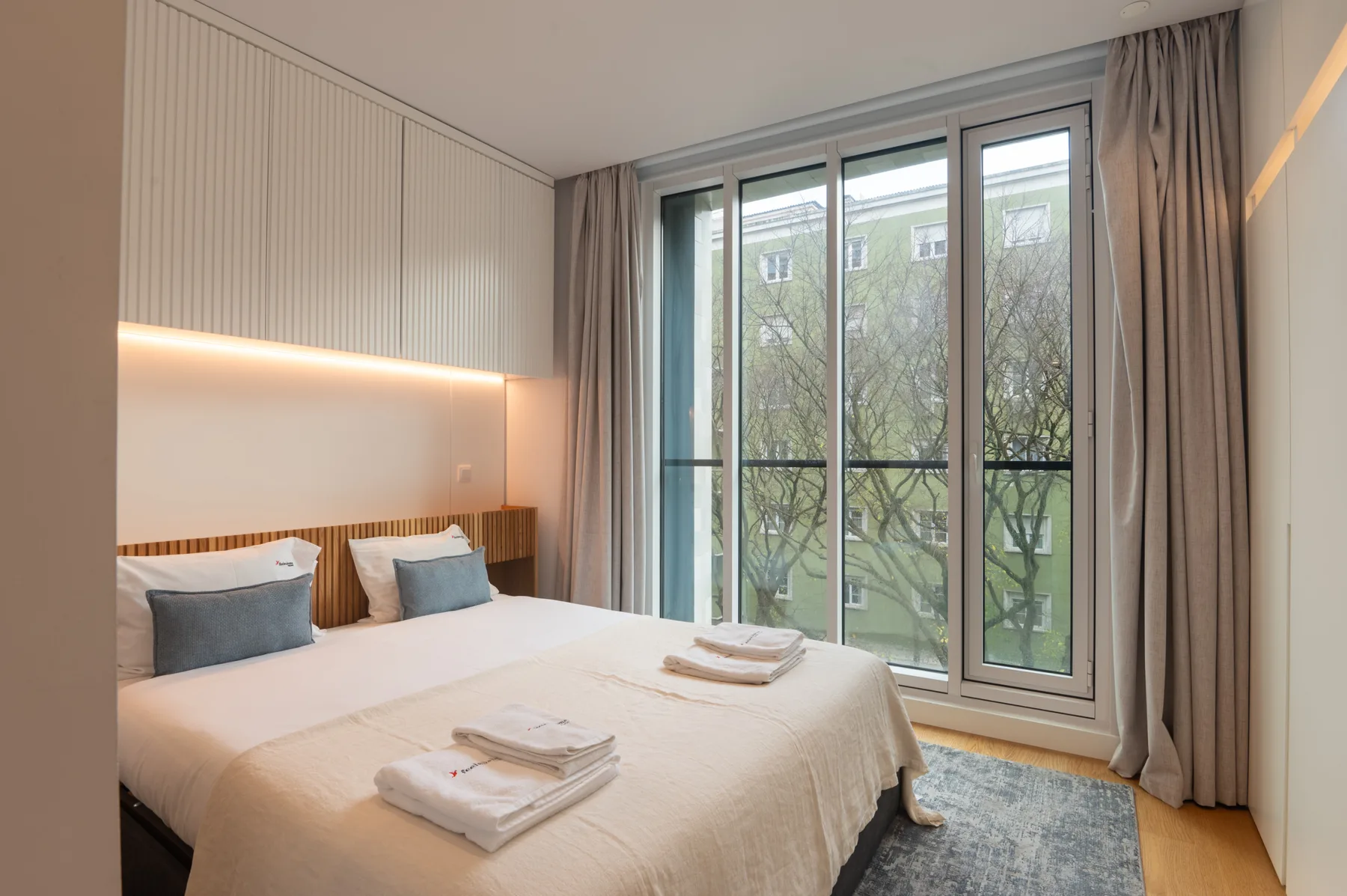 A bedroom with a neatly made bed beside tall windows and curtains.