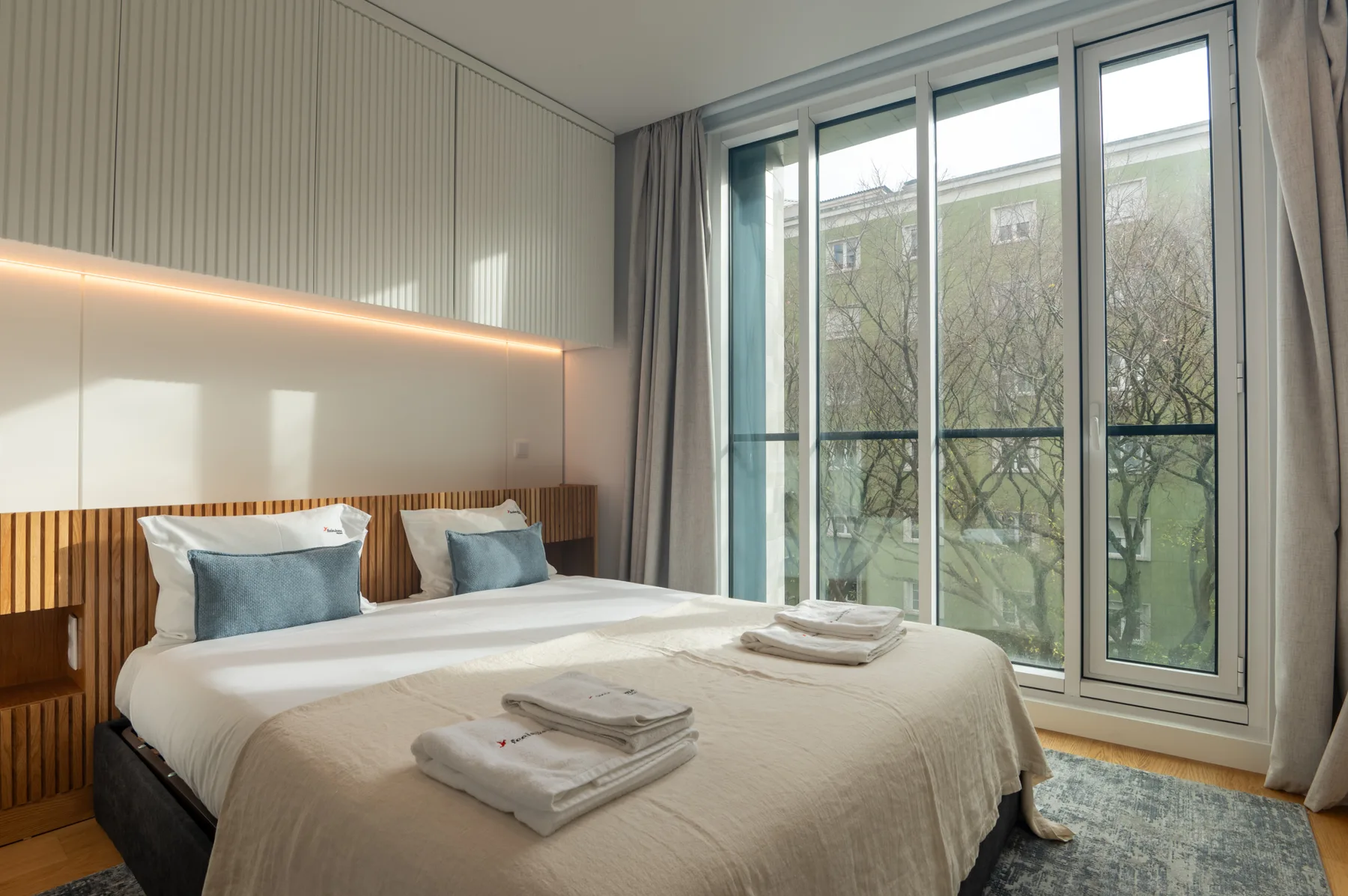 A bedroom with a neatly made bed beside large windows.