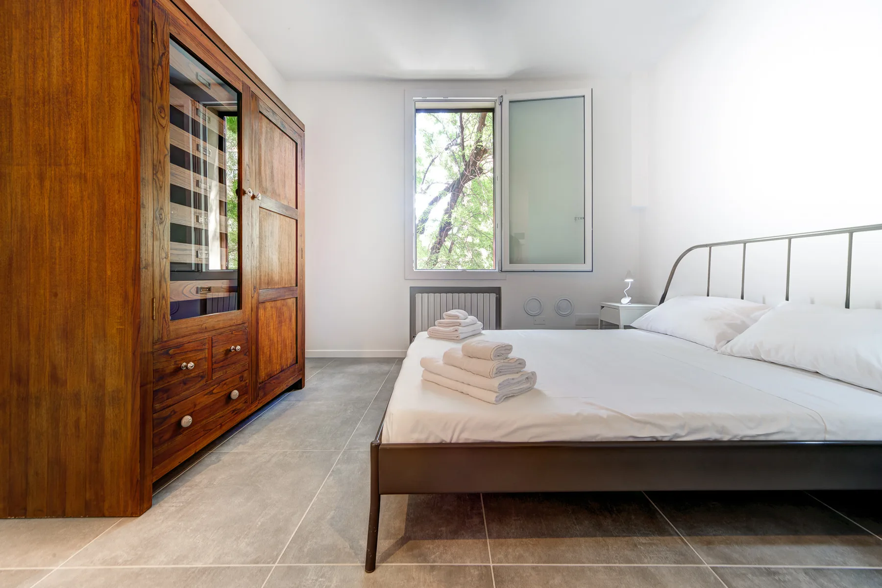Un dormitorio luminoso con una cama junto a un armario de madera y una ventana abierta.