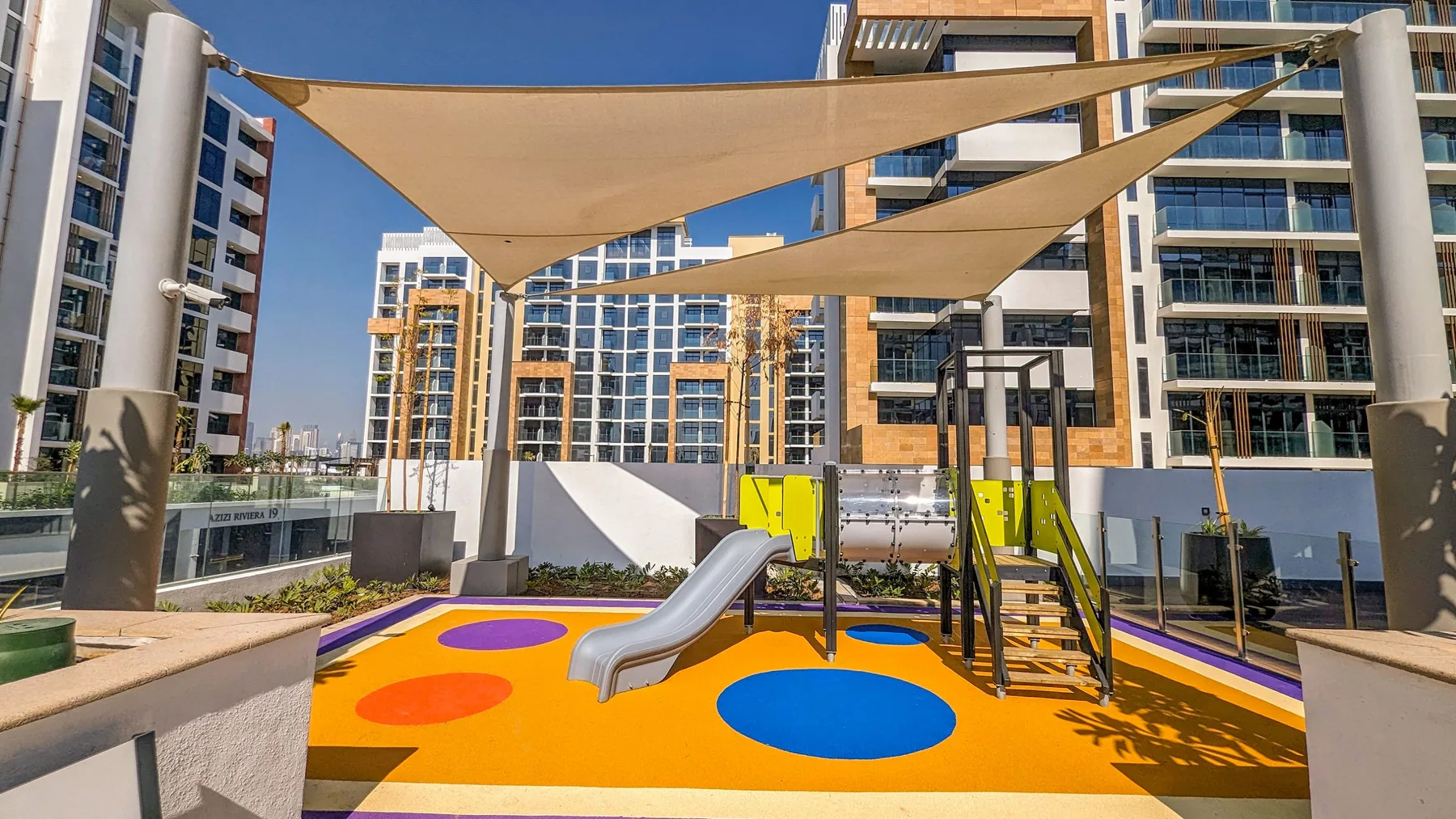 Un parque infantil al aire libre y colorido, con toboganes y toldos de sombra entre edificios.