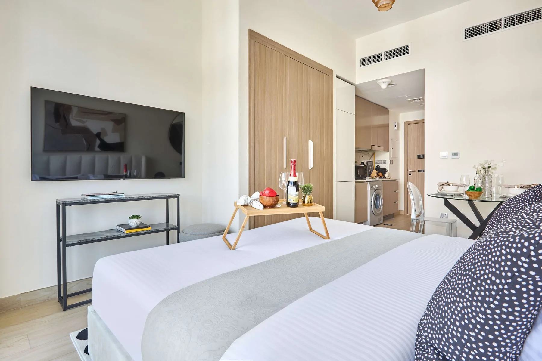 Un dormitorio con una cama, un televisor en la pared y una pequeña cocina al fondo.