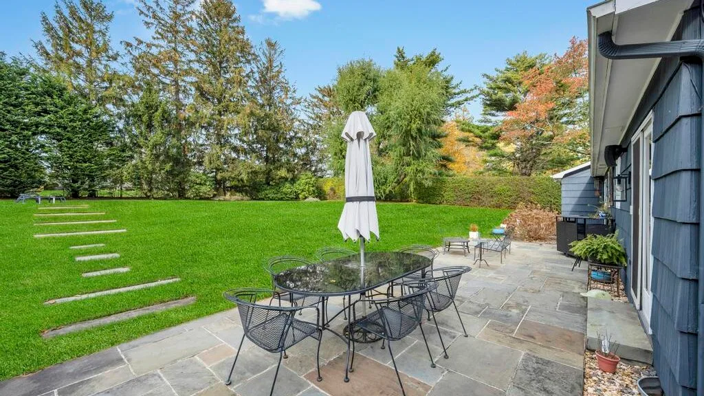 A stone patio with outdoor seating overlooks a green lawn and leafy trees.