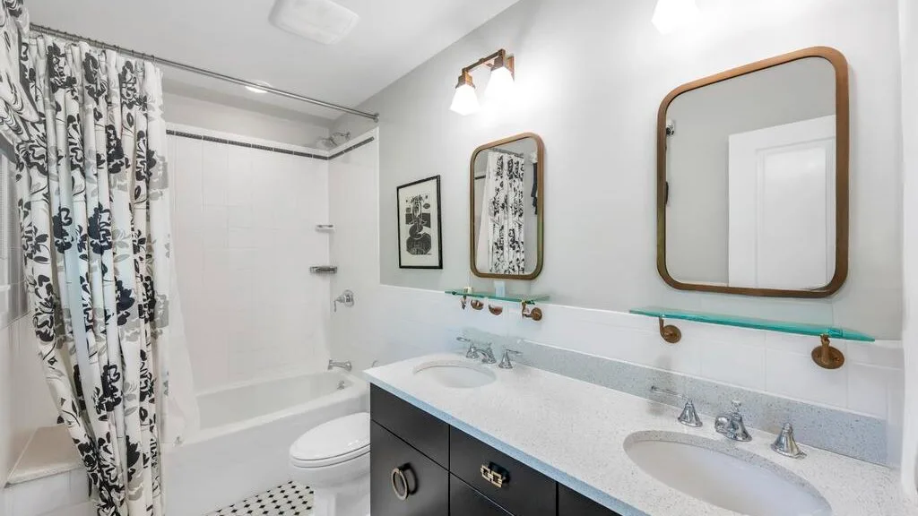 A bathroom with a long vanity, two sinks, and a bathtub behind a patterned shower curtain.