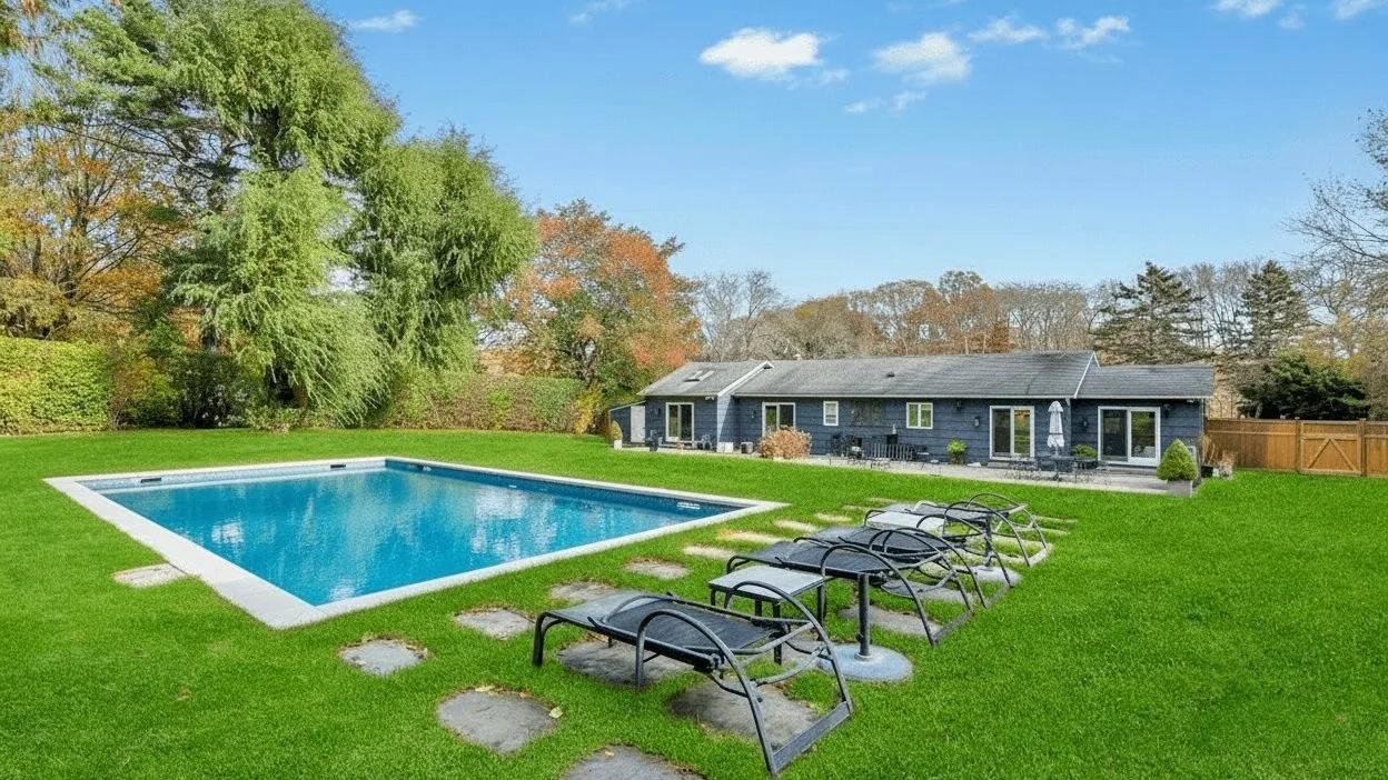 A backyard pool beside a lawn, with lounge chairs and a house in the background.