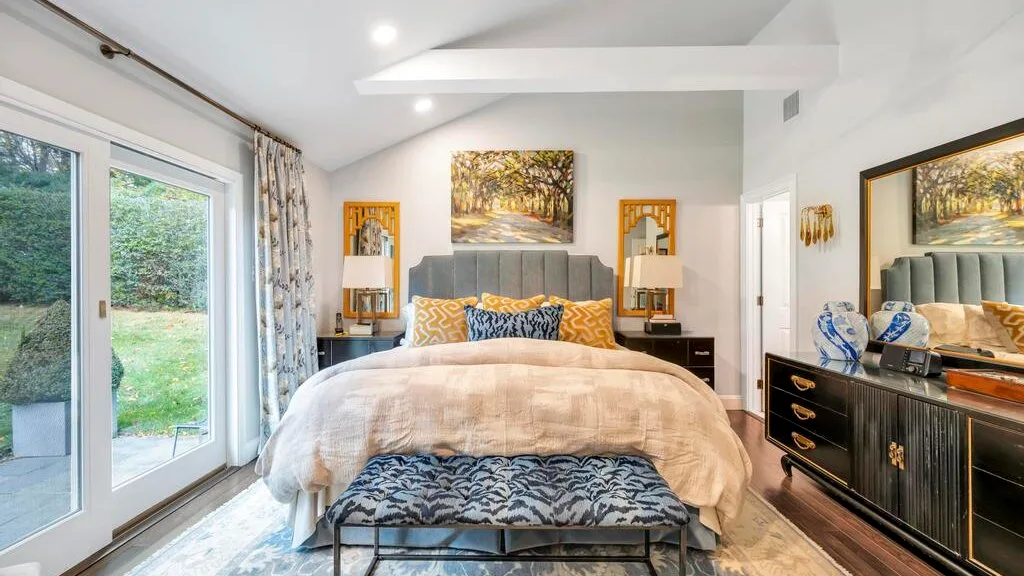 A bright bedroom with a bed, patterned bench, and large glass doors.
